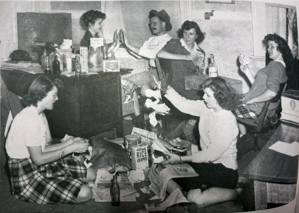 A black and white photo of a cluttered room with several women talking, playing cards, and organizing papers.